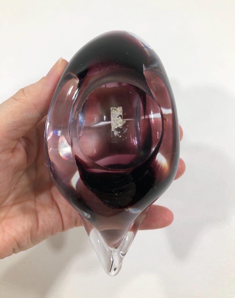 May include: A hand is holding a clear glass vase with a deep purple interior and a clear, pointed base. The vase has a rounded top and a smooth, polished surface. The color transitions from clear to deep purple.