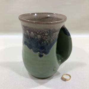 May include: A handmade ceramic mug with a unique glaze. The mug features a sage green base, transitioning to a dark blue and brown rim. A gold ring with three small diamonds sits beside the mug.