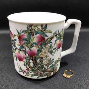 May include: White ceramic mug with a peach tree design. The mug has a handle and is decorated with pink peaches, green leaves, and white flowers.