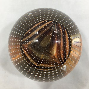 May include: A clear glass paperweight with a swirling brown and white pattern inside. The glass is covered in small air bubbles.