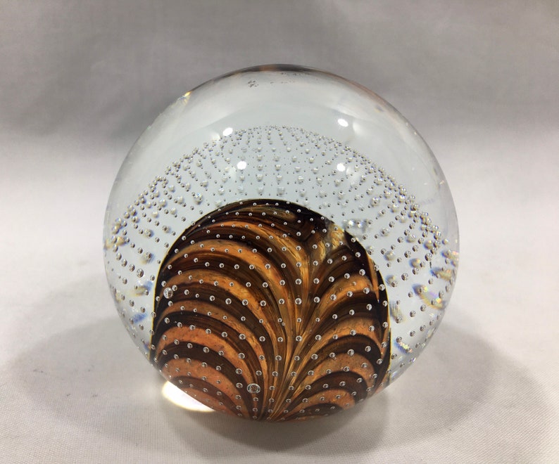 May include: A clear glass paperweight with a swirling brown and gold design inside. The paperweight is filled with small air bubbles.