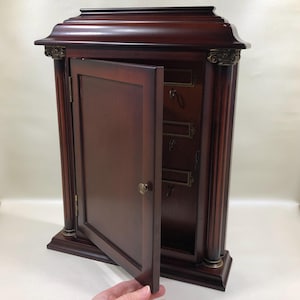 May include: A dark cherry-stained wooden key cabinet with an open door. The cabinet has a classic design with decorative columns and brass accents. The interior features key hooks and label holders.