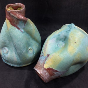 May include: Two ceramic vases with a unique, abstract, and organic shape. The vases are glazed in shades of green, yellow, and brown.