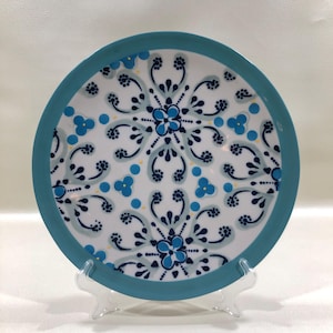 May include: A round ceramic plate with a white background and a teal border. The plate features a symmetrical floral design in shades of blue and teal, with small yellow accents. The plate is displayed on a clear plastic stand.