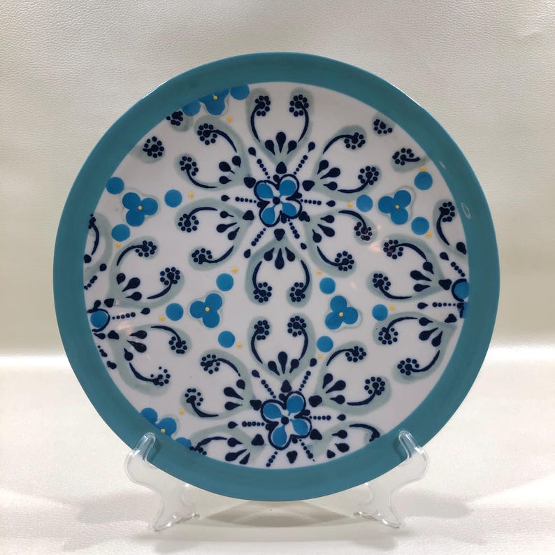 May include: A round ceramic plate with a white background and a teal border. The plate features a symmetrical floral design in shades of blue and teal, with small yellow accents. The plate is displayed on a clear plastic stand.