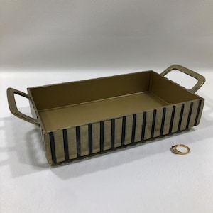 May include: A rectangular gold-toned metal tray featuring a ribbed exterior with vertical black accents and two integrated handles on the sides. A small gold ring with a clear stone is positioned on the surface next to the tray for scale.