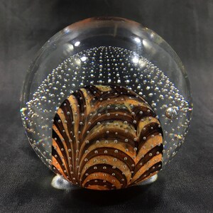 May include: A clear glass paperweight with a swirling pattern of brown and gold inside. The paperweight is filled with small air bubbles.