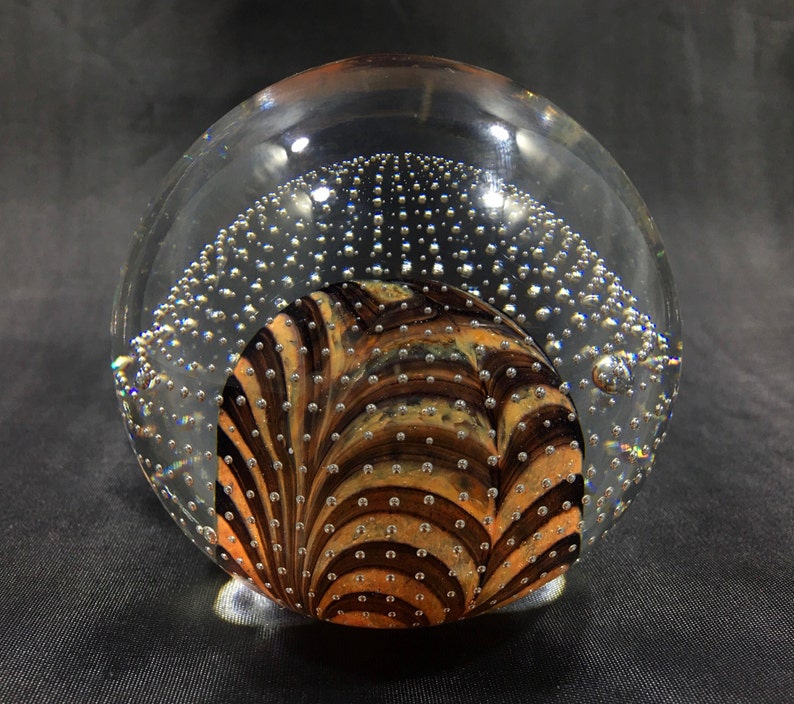 May include: A clear glass paperweight with a swirling pattern of brown and gold inside. The paperweight is filled with small air bubbles.