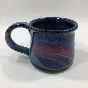 May include: A handmade ceramic mug with a glossy finish. The mug features a dark blue glaze with streaks of purple, brown, and teal. It has a rounded body, a sturdy handle, and a slightly flared rim. A unique piece for enjoying coffee or tea.