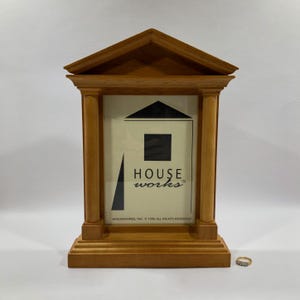 May include: Wooden picture frame with a pediment top and columns. The frame has a light brown finish and a rectangular opening. The text "HOUSE works" is visible on the cream-colored insert. A small gold ring with clear stones sits in front of the frame.