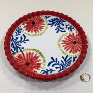 May include: A round, white ceramic plate with a red, scalloped edge. The plate features red and blue floral designs. A gold ring with small stones is next to the plate.