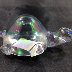 May include: A clear iridescent glass turtle figurine with a rounded shell and a long neck. The turtle is made of glass and has a shimmering, iridescent finish.