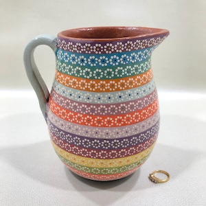 May include: A colorful ceramic pitcher with a rainbow striped design. Each stripe features a pattern of small white dots and blue dots. The handle is light blue, and the rim is brown. A gold ring with three small stones sits next to the pitcher.