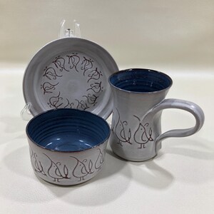 May include: A ceramic tableware set featuring a bowl, a mug, and a plate. The set is primarily gray with blue interiors and brown bird-like designs. The mug has a curved handle, and the plate has a decorative rim.