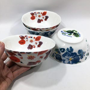 May include: A set of four white ceramic bowls. The bowls feature a floral design with red, purple, and white flowers. The underside of one bowl has a blue floral design and text. The bowls are suitable for serving food.