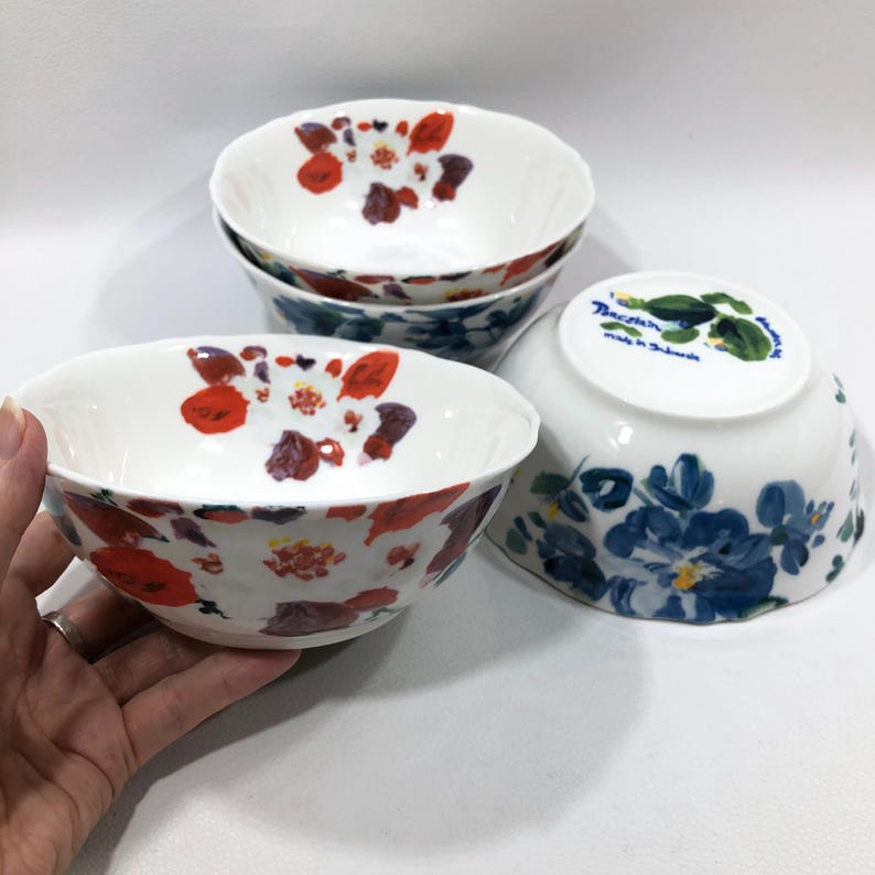 May include: A set of four white ceramic bowls. The bowls feature a floral design with red, purple, and white flowers. The underside of one bowl has a blue floral design and text. The bowls are suitable for serving food.