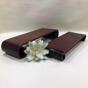 May include: Two rectangular, dark red-brown wooden display stands with curved ends. A white and green flower rests between the stands. The stands are likely for displaying small items.