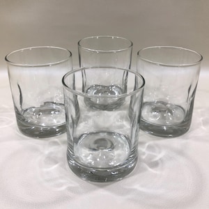 May include: Four clear, short, round drinking glasses. The glasses have a thick base and a slightly textured appearance. The glasses are empty and appear to be made of glass. The glasses are arranged in a group on a white surface.