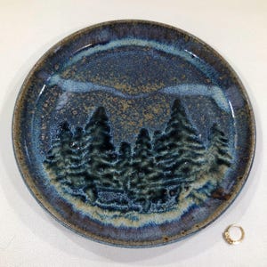 May include: A round ceramic plate with a forest scene. The plate features a dark blue glaze with a textured appearance. The forest is depicted with dark green trees. The rim has a brown and tan color.