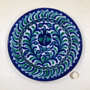 May include: Round ceramic plate with a blue and green floral design. The plate has a white background and a gold ring is next to it. The plate's rim is dark blue.