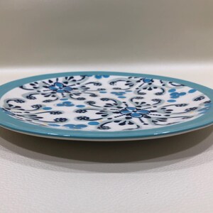 May include: A round ceramic plate with a white center featuring a blue floral pattern. The rim of the plate is a solid light blue. The plate is on a white surface.