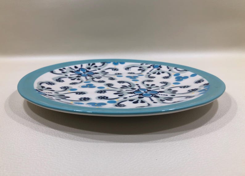 May include: A round ceramic plate with a white center featuring a blue floral pattern. The rim of the plate is a solid light blue. The plate is on a white surface.
