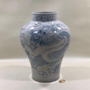 May include: A white ceramic vase with a blue hand-painted dragon motif wrapping around the body. The vase features a wide, rounded shoulder, a narrow neck, and a flared rim. A small gold ring is placed on the surface next to the base for scale.