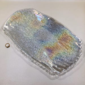 May include: Iridescent glass tray with a textured surface, displaying a spectrum of colors. A small gold ring with a gemstone is next to the tray. The tray's shape is rectangular with irregular edges, suitable for home decor.