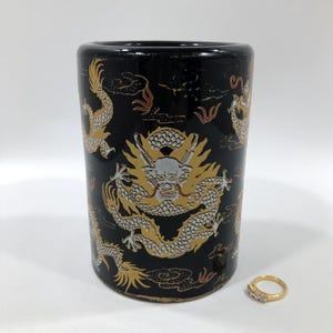 May include: Black ceramic cylindrical vase with gold and white dragon designs. The vase has a glossy finish and a small gold ring with clear stones is next to it.