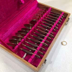 May include: A vintage flatware set in a wooden box with a bright pink velvet lining. The set includes spoons, forks, and knives with gold-toned metal and dark brown handles. A gold ring with small stones sits to the side.