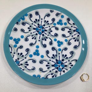 May include: A round, white ceramic plate with a turquoise rim. The plate features a repeating pattern of blue floral designs and dots. A gold ring with small stones is next to the plate.