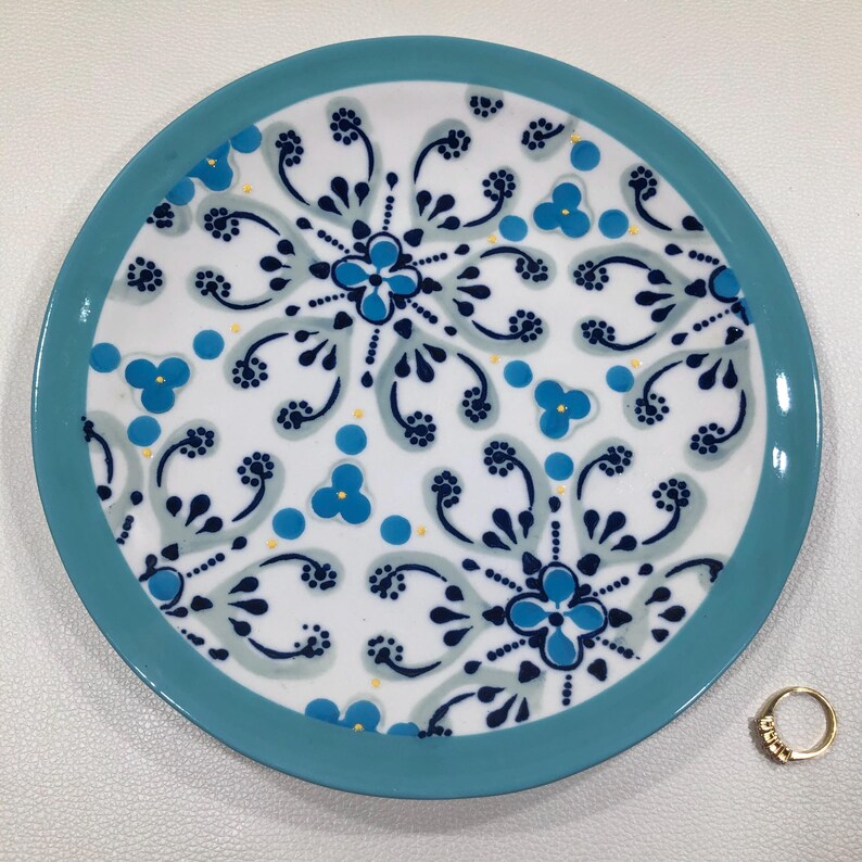 May include: A round, white ceramic plate with a turquoise rim. The plate features a repeating pattern of blue floral designs and dots. A gold ring with small stones is next to the plate.