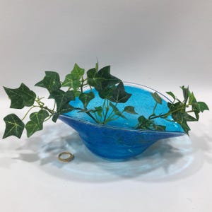 May include: A blue glass bowl with a textured surface, filled with green ivy. A small gold ring with clear stones sits near the bowl. The bowl has a white rim and a light blue interior.