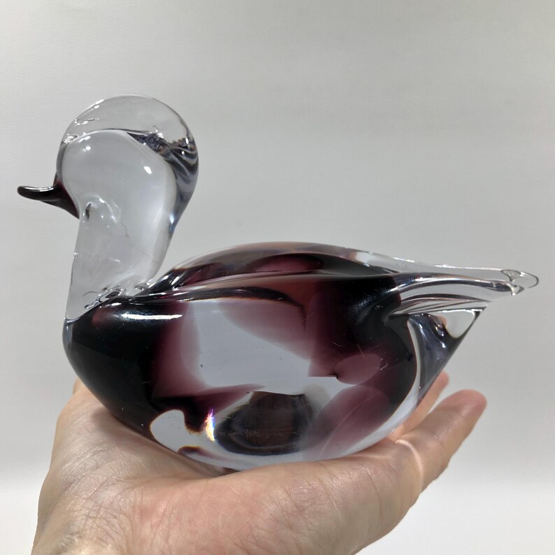 May include: A clear and purple glass duck figurine. The duck has a long neck and a small black beak. The body of the duck is a mix of clear and purple glass. The figurine is held in a hand.