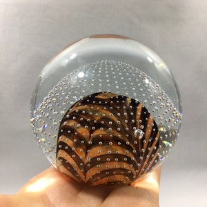May include: A clear glass paperweight with a swirling pattern of brown and orange inside. The glass is filled with small air bubbles.