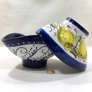 May include: Two ceramic bowls with a white and blue design. One bowl is upright, the other is tilted, revealing a yellow fruit design. A gold ring with a small stone sits in front of the bowls.
