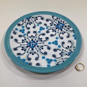 May include: A round, white ceramic plate with a turquoise rim and intricate blue floral patterns. A gold ring with small stones sits to the side of the plate. The plate's design features a symmetrical arrangement of stylized flowers and leaves.