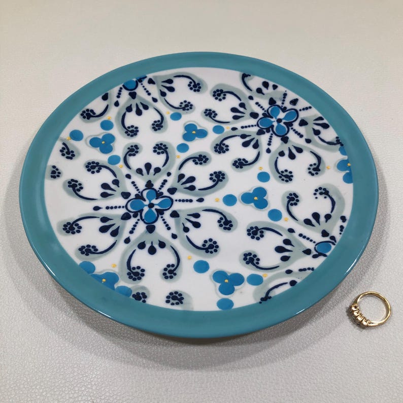 May include: A round, white ceramic plate with a turquoise rim and intricate blue floral patterns. A gold ring with small stones sits to the side of the plate. The plate's design features a symmetrical arrangement of stylized flowers and leaves.