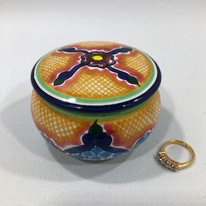 May include: A round, hand-painted ceramic trinket box with a lid, featuring a vibrant orange background and colorful floral designs in blue, red, and green. A gold ring with small diamonds sits beside the box.