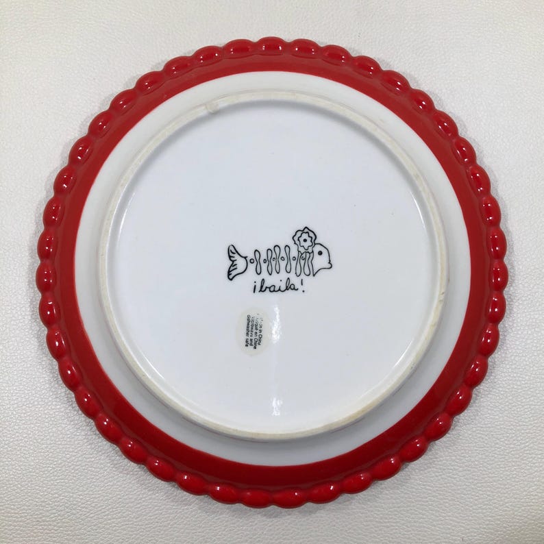 May include: A white ceramic plate with a red scalloped edge. The plate features a fish skeleton design and the text "i baila!". The plate is likely for serving food.