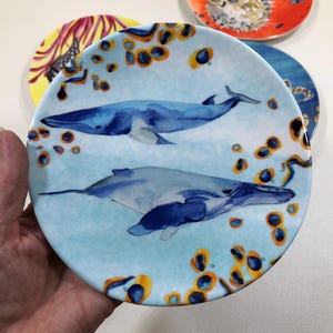 Może przedstawiać: Okrągły ceramiczny talerz z akwarelową ilustracją dw&oacute;ch płetwali błękitnych pływających w jasnoniebieskiej wodzie, otoczonych rozproszonymi pomarańczowymi i ciemnoniebieskimi kolistymi wzorami, kt&oacute;re pokrywają całą powierzchnię talerza.
