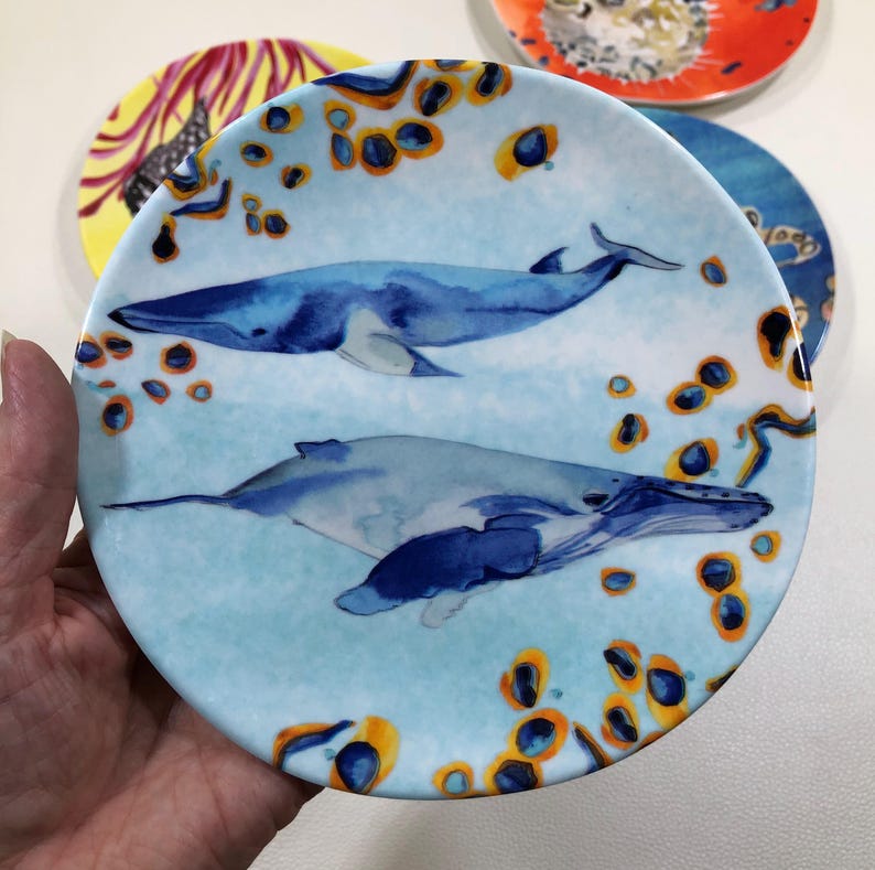 Może przedstawiać: Okrągły ceramiczny talerz z akwarelową ilustracją dw&oacute;ch płetwali błękitnych pływających w jasnoniebieskiej wodzie, otoczonych rozproszonymi pomarańczowymi i ciemnoniebieskimi kolistymi wzorami, kt&oacute;re pokrywają całą powierzchnię talerza.