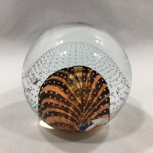 May include: A clear glass paperweight with a swirling pattern of brown and orange stripes and small air bubbles throughout.