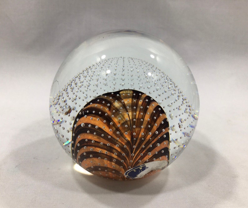 May include: A clear glass paperweight with a swirling pattern of brown and orange stripes and small air bubbles throughout.