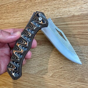 May include: A folding pocket knife with a patterned handle in black, white, and bronze tones. The blade is silver and partially open, revealing a sharp edge. The knife is held against a wooden surface.