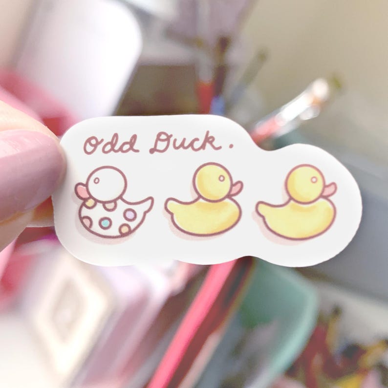 Puede incluir: Una pegatina con el texto "Odd Duck." La pegatina presenta tres patos de dibujos animados. Un pato es amarillo con un pico y patas rosas. Los otros dos patos son amarillos con picos y patas rosas. Uno de los patos amarillos tiene lunares blancos.