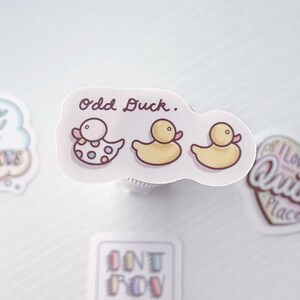 Puede incluir: Una pegatina con el texto "Odd Duck."  Se representan tres patos de dibujos animados en la pegatina. Un pato es blanco con lunares rosas y azules. Los otros dos patos son amarillos con picos y patas rosas.