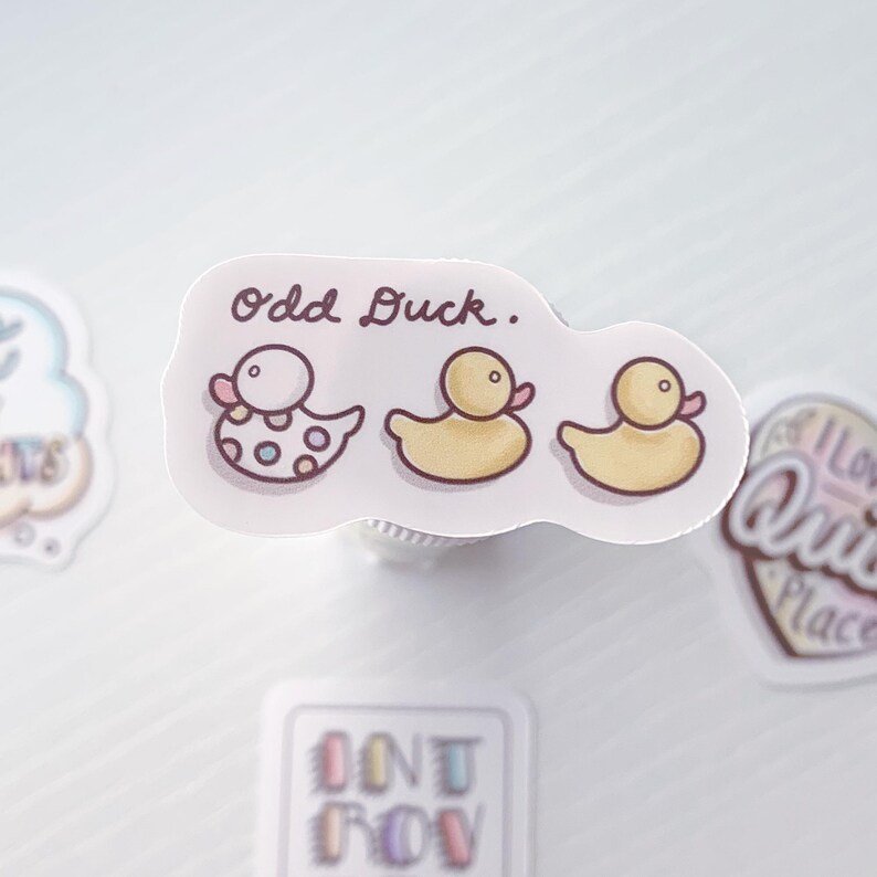 Puede incluir: Una pegatina con el texto "Odd Duck."  Se representan tres patos de dibujos animados en la pegatina. Un pato es blanco con lunares rosas y azules. Los otros dos patos son amarillos con picos y patas rosas.