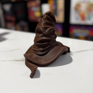 May include: A brown, felt-like, wizard's hat with a pointed top and a wide brim. The hat is made of a soft, pliable material and has a slightly wrinkled texture.