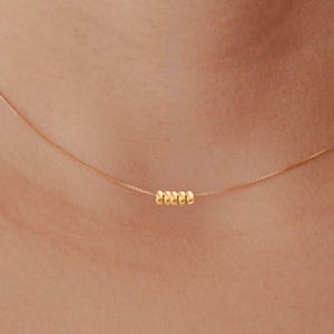 Könnte beinhalten: Zarte goldene Halskette mit einer feinen Kette und einer kleinen Gruppe runder Perlen. Die Halskette wird um den Hals getragen und unterstreicht ihr minimalistisches Design und ihre dezente Eleganz. Die warme Goldfarbe kontrastiert mit dem Hautton.